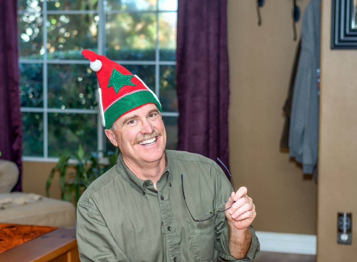 Senior male holding glasses wearing a holiday elf hat at home