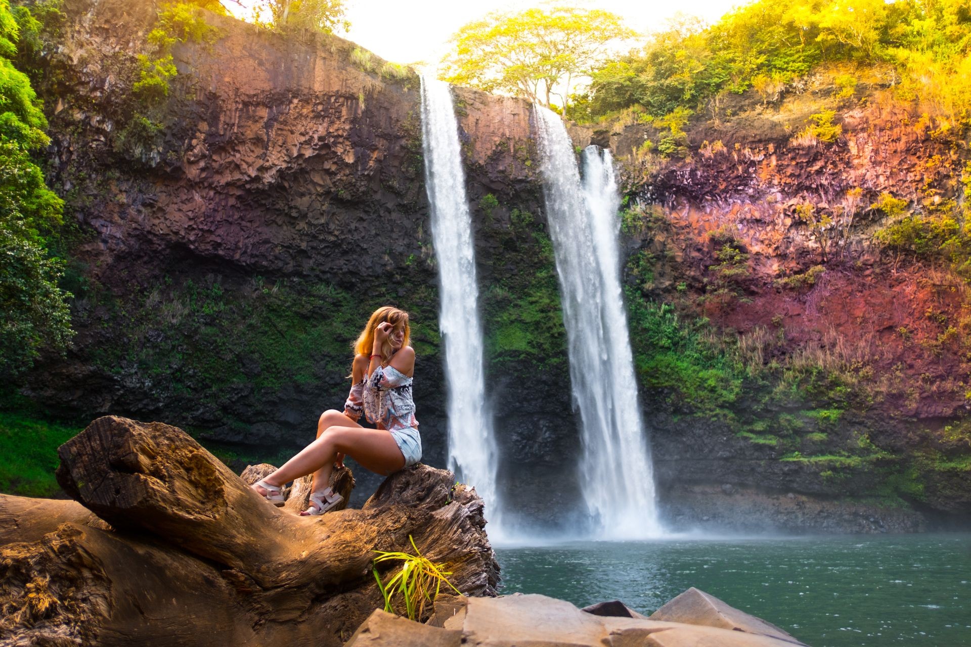 alone traveler woman seats with her back at stone and looks at colorful Hawaiian waterfall. hawaii. traveling