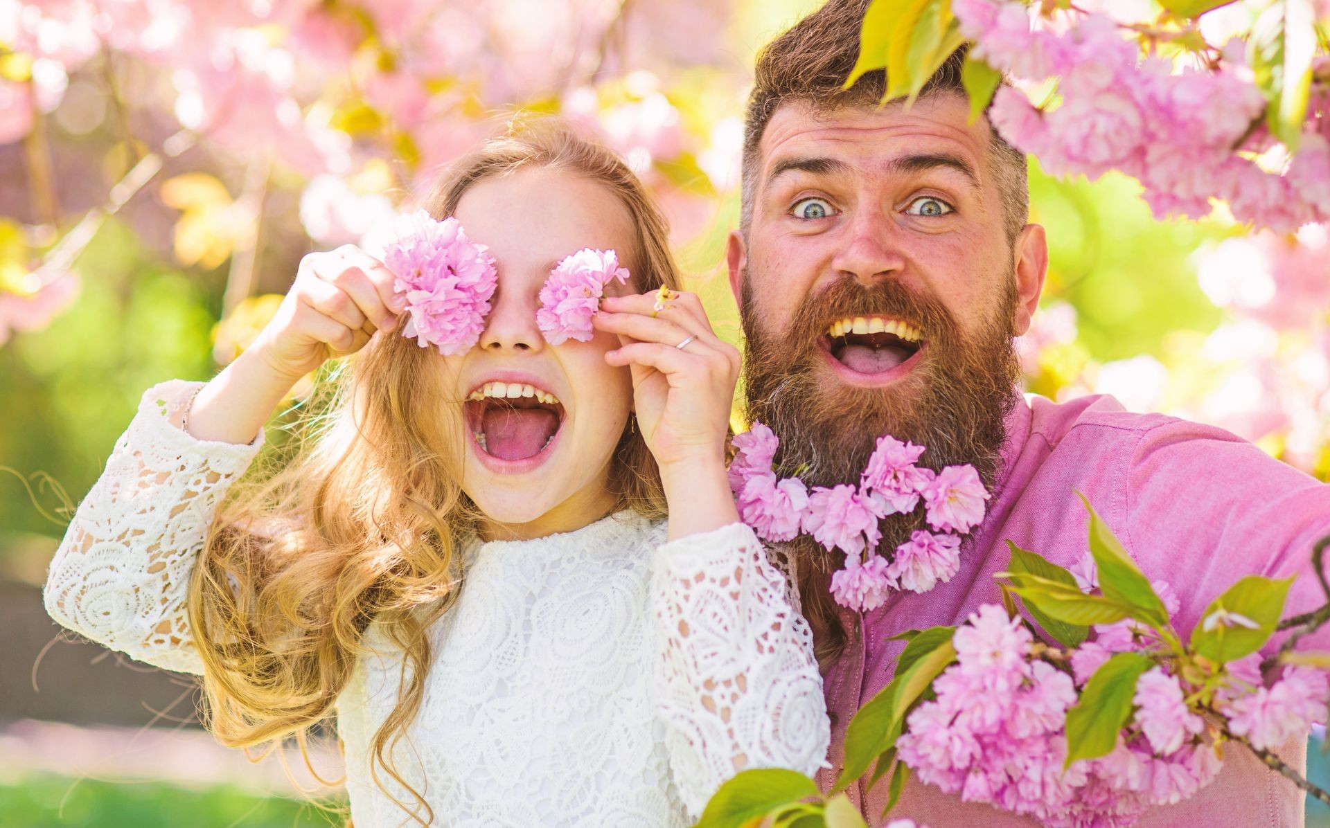 Child and man with tender pink flowers in beard. Father and daughter on happy face play with flowers as glasses, sakura background. Girl with dad near sakura flowers on spring day. Family time concept