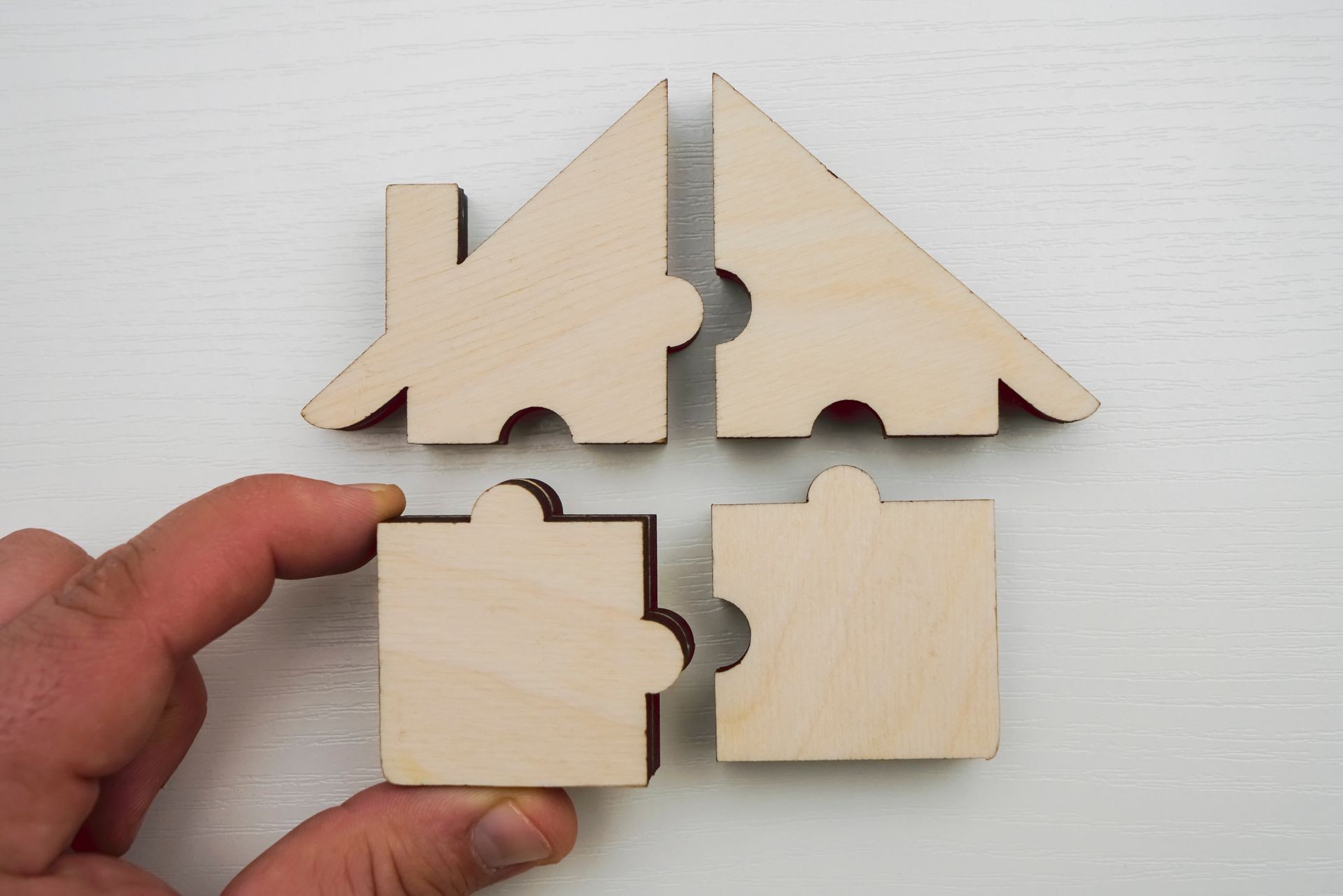  wooden house made from four pieces - puzzles on white wooden background. one jigsaw between two male fingers. teamwork idea. 