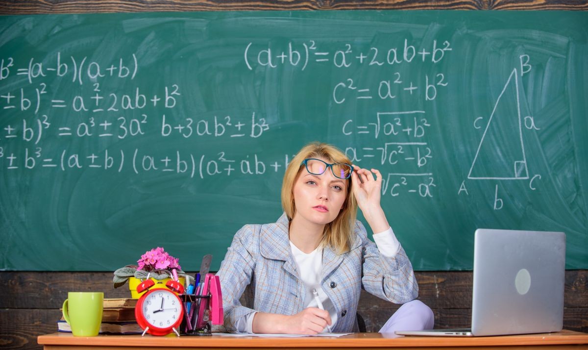 Teacher doubtful face listening you. She has doubts in your report or knowledge. Teacher woman sit table classroom chalkboard background. Teacher suspiciously peeking out of eyeglasses.