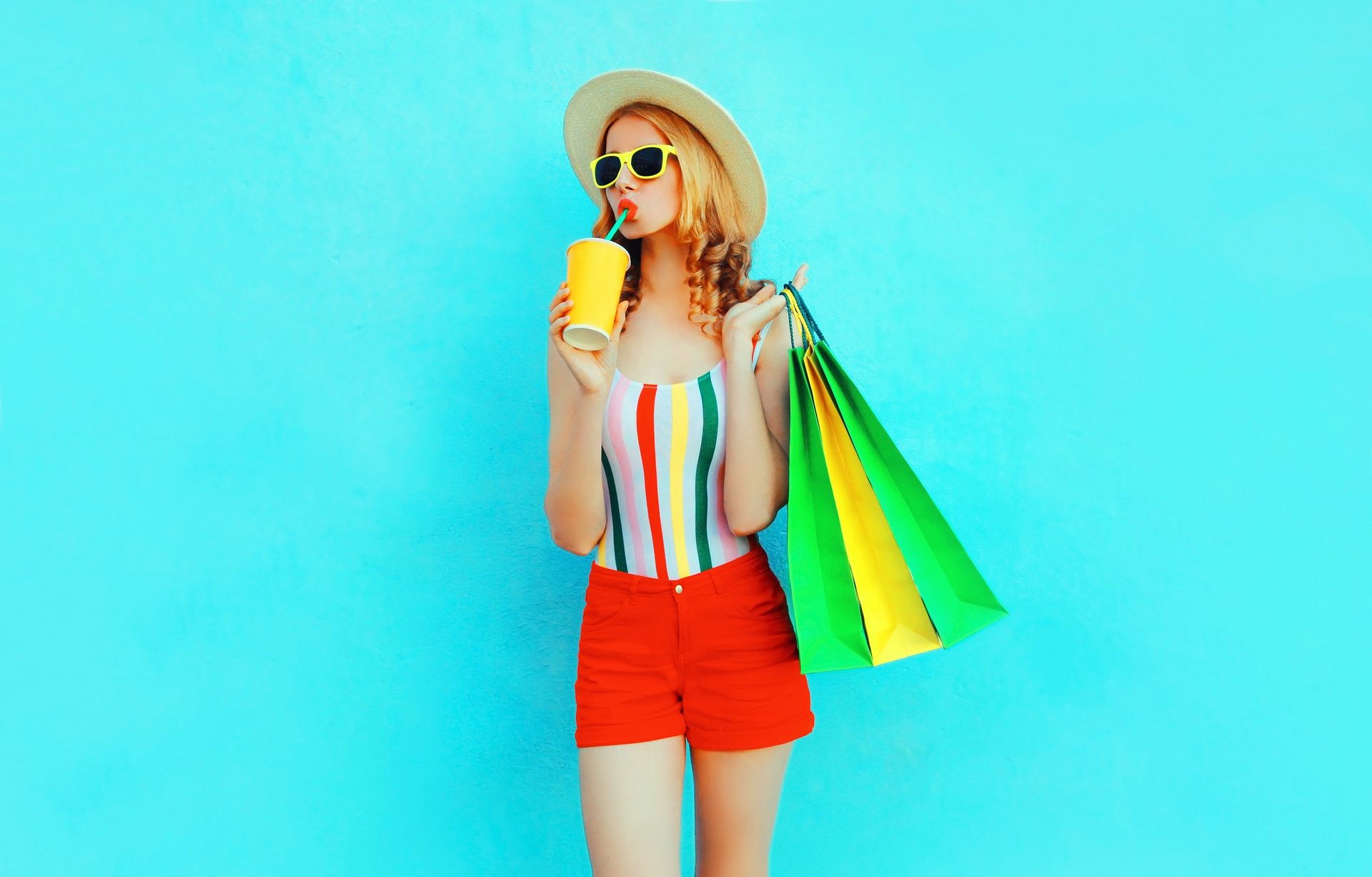 Young woman drinking juice holding shopping bags in colorful t-shirt, summer straw hat, sunglasses, red shorts on blue background