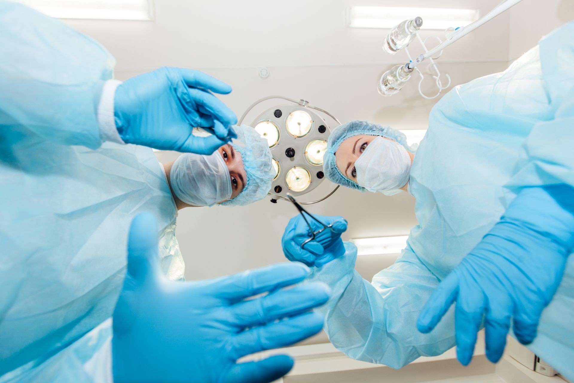 Two surgeons in protective uniform preparing for operation, looking at camera on background of surgical lamp. 