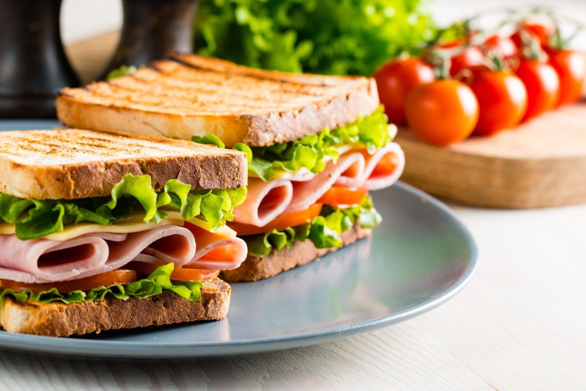 Close-up of two sandwiches with bacon, salami, prosciutto and fresh vegetables on rustic wooden cutting board. Club sandwich concept.