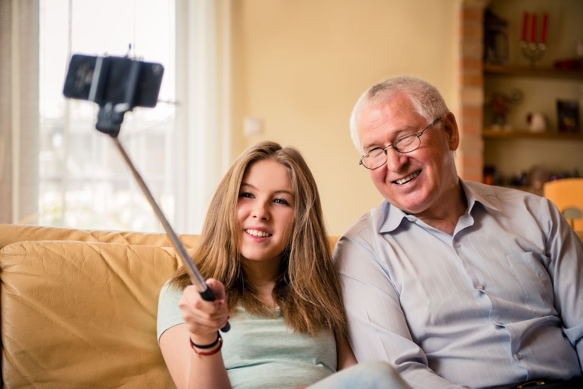 Teenage girl taking photo with mobile phone on selfie stick of herself and her grandfather at home