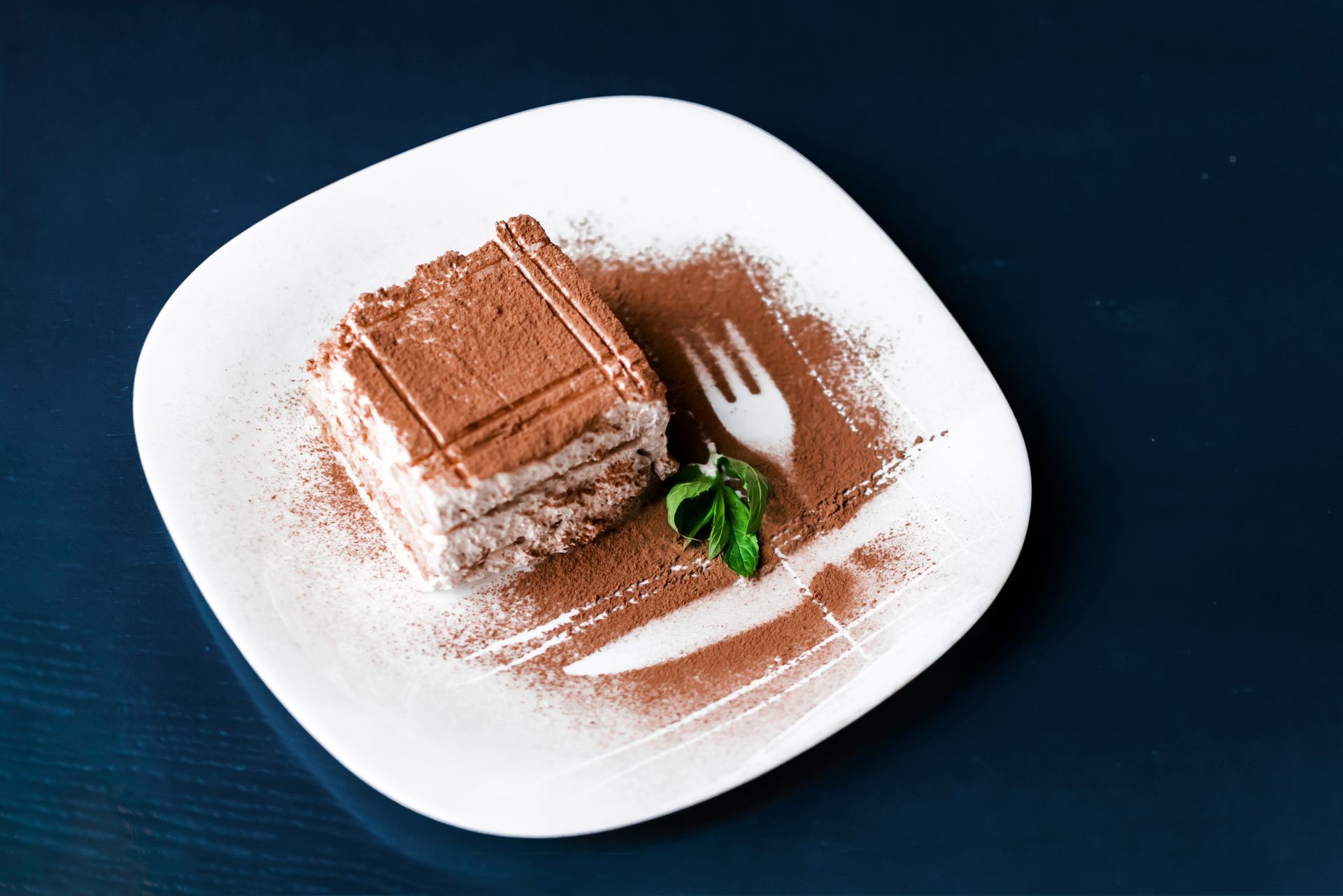 Tiramisu traditional Italian dessert on white plate on dark blue table. Close up top view food with fork and knife decor