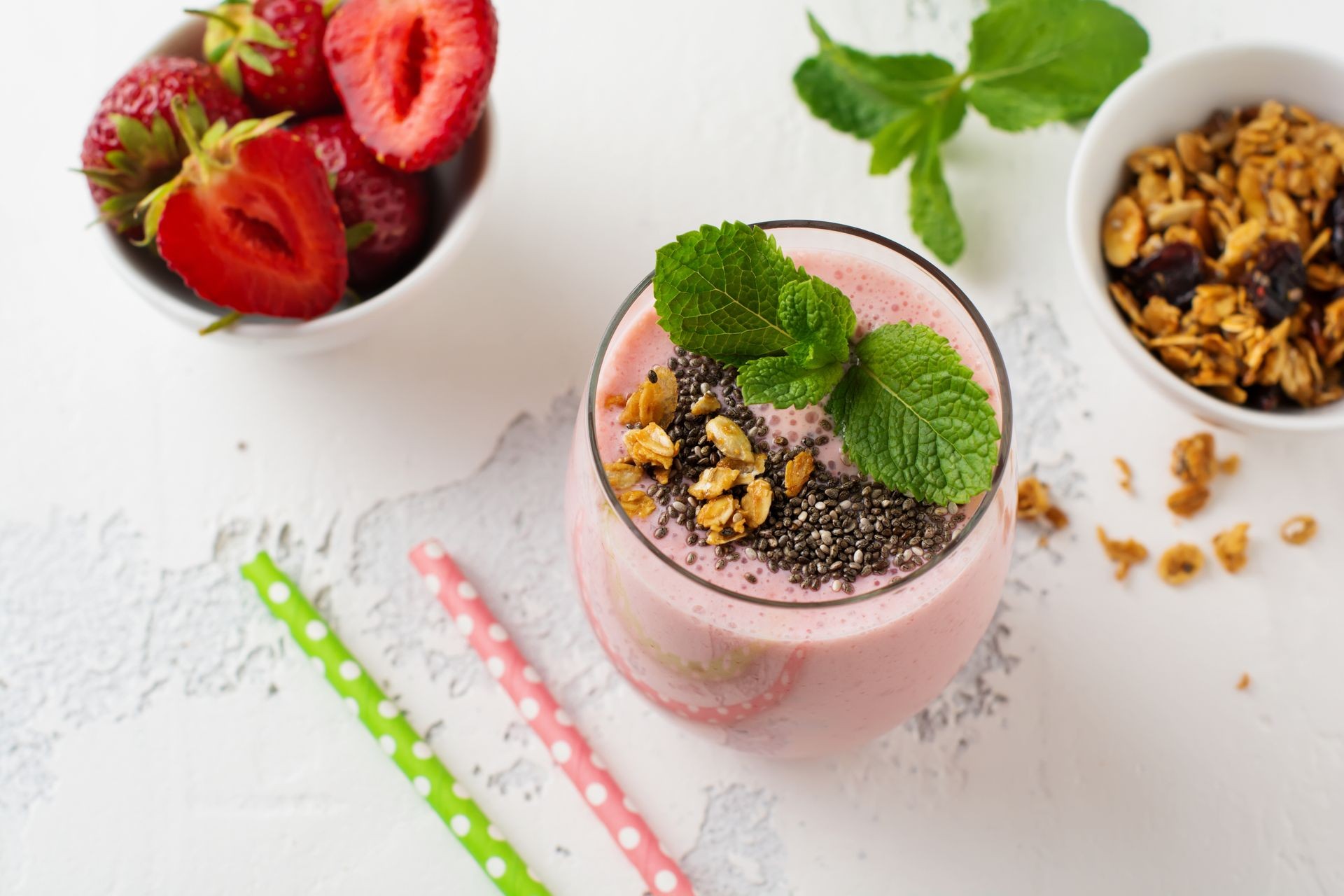 Pink strawberries, banana smoothie with granola and chia seeds in glass on light white concrete background. Top view. Copy space.