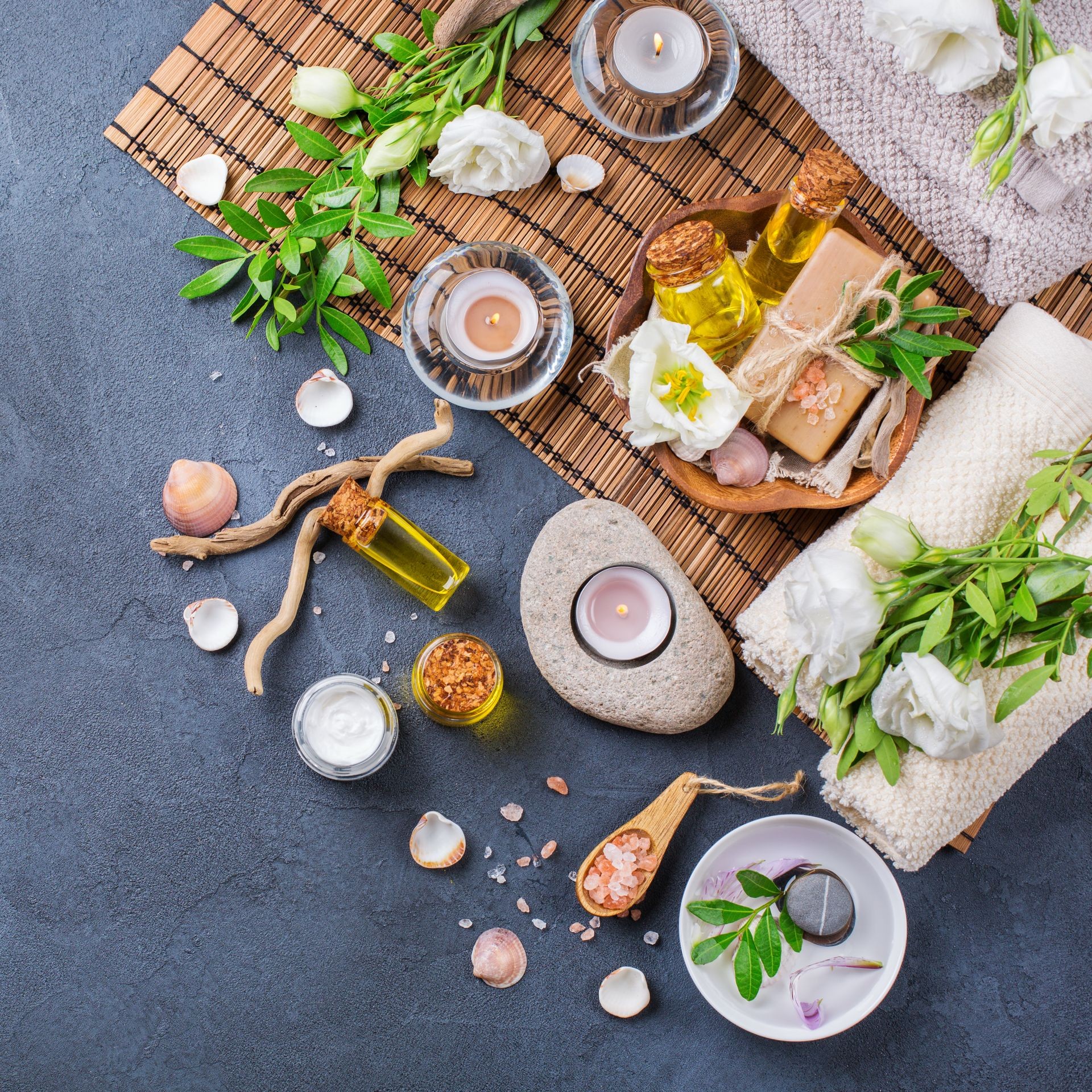 Spa still life wellness setting concept. Assortment of essential oil cream sea salt natural soap candles white spring flowers and towel on a black table. Top view, flat lay